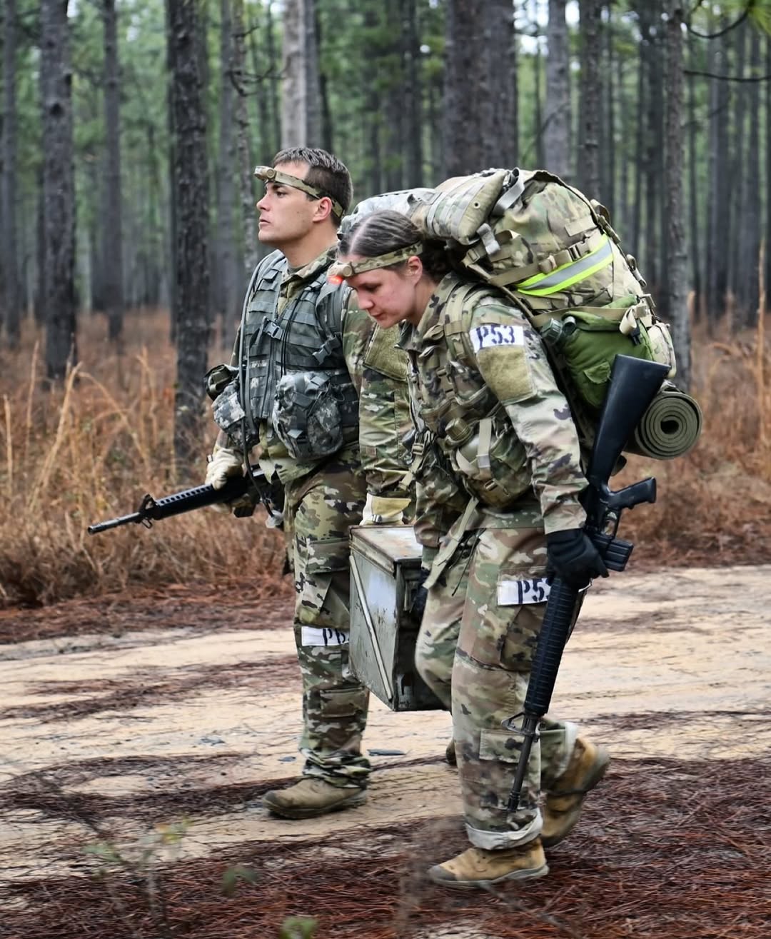 Two soldiers in camouflage uniforms carry heavy backpacks while walking through a forest. The setting is rugged, conveying determination and teamwork.