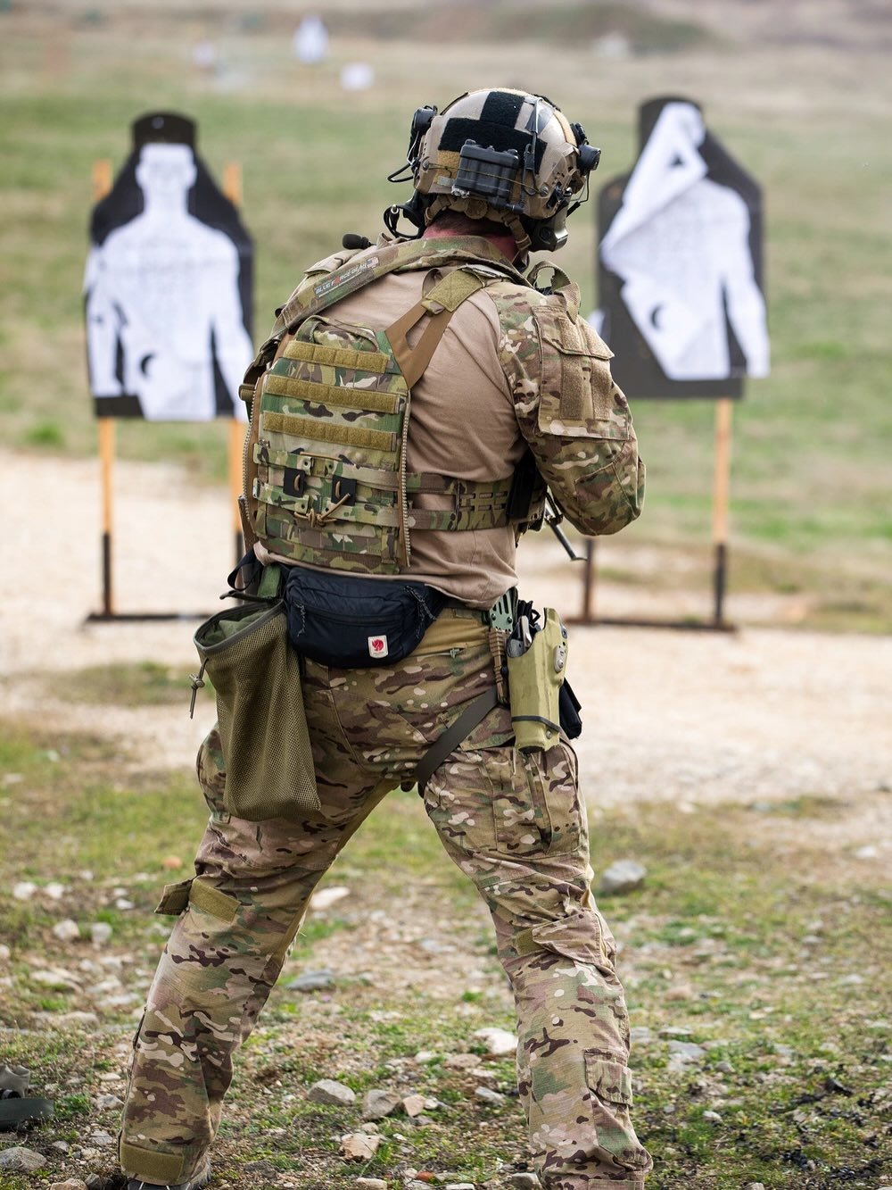 A soldier in camouflage and tactical gear aims a rifle at two silhouette targets during a training exercise on a grassy field.