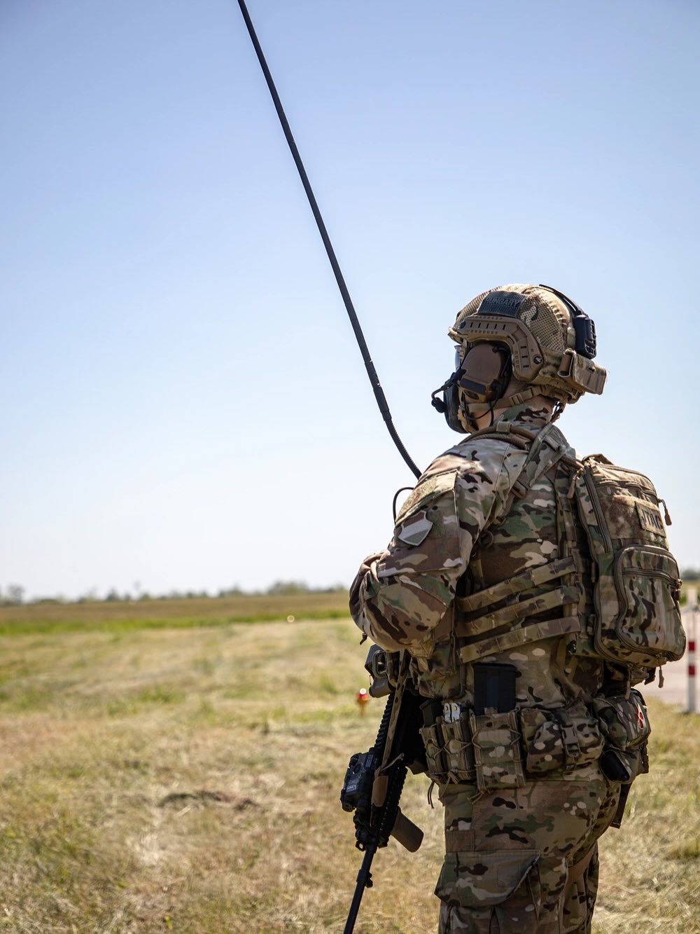 A soldier in camouflage gear stands on grassy terrain, holding a tall antenna. The blue sky and open field emphasize readiness and communication.