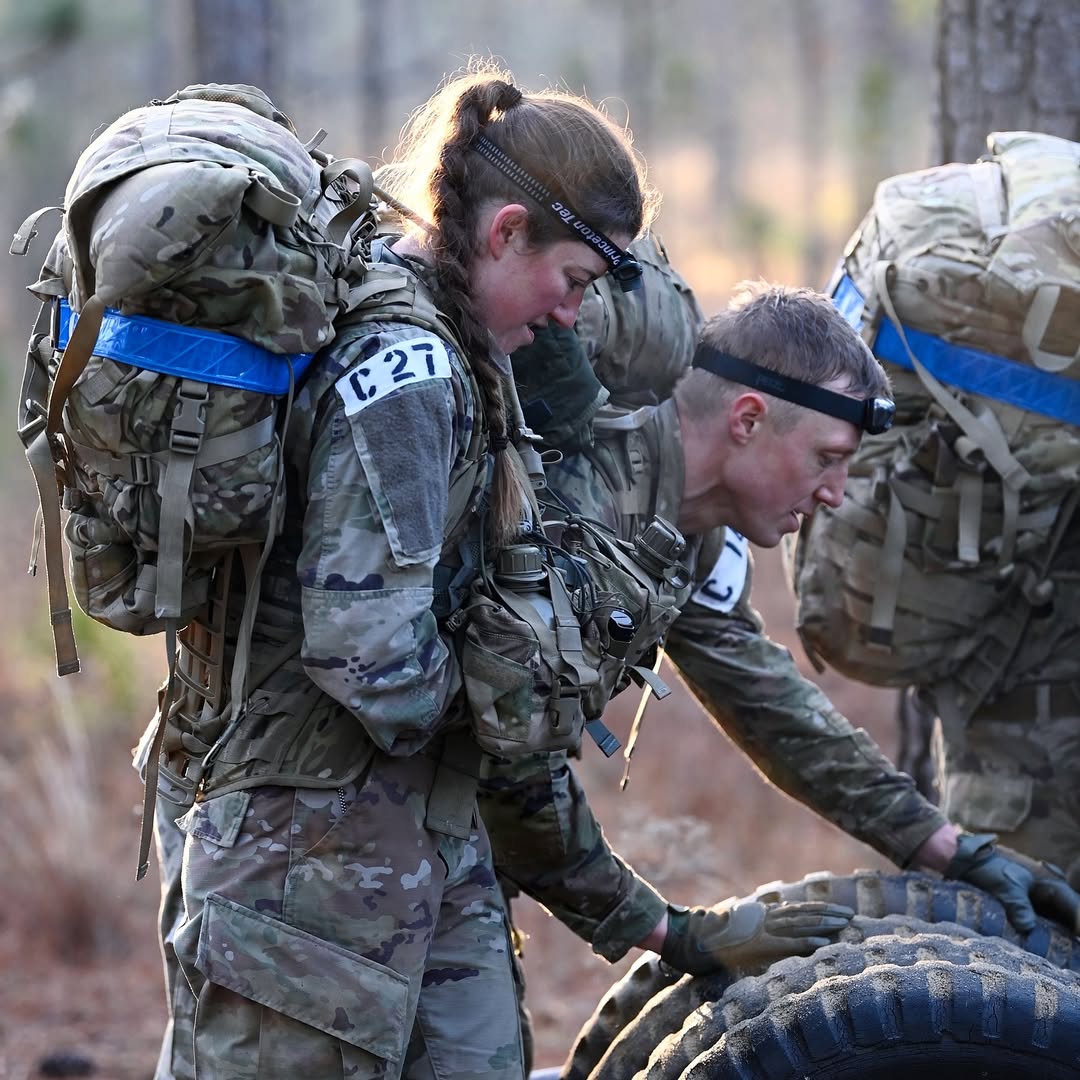 Soldiers in camouflage gear carry large backpacks through a wooded area, focusing on teamwork and endurance. The scene conveys determination.