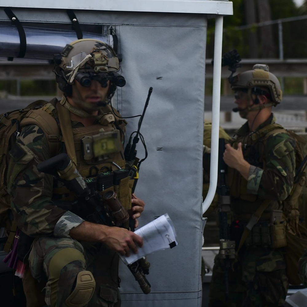 Two soldiers in camouflage uniforms, holding rifles, stand by a covered structure. The setting appears tense, suggesting a military operation.