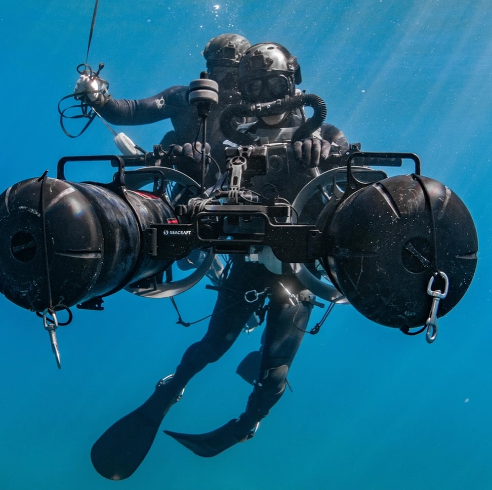 Diver underwater equipped with advanced gear, holding a propulsion device. Sunlight filters through the water, creating a serene, exploratory atmosphere.