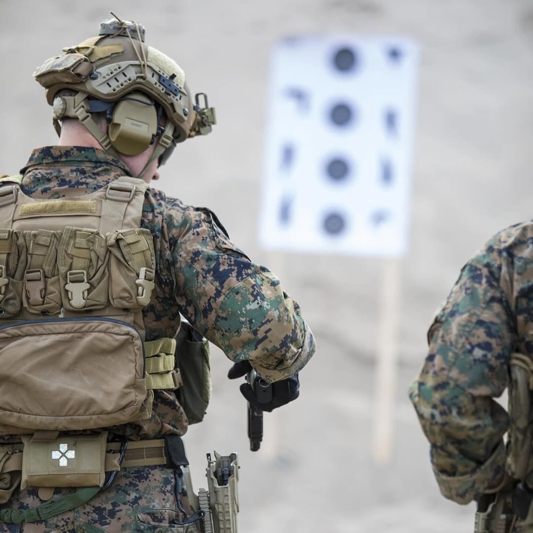 Two soldiers in camouflage gear and helmets face a blurred target range, creating a focused and intense training atmosphere.