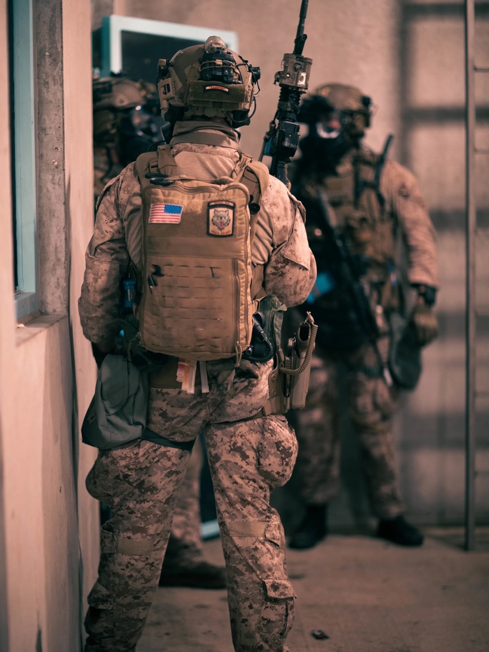 Armed soldiers in tactical gear stand in formation indoors, focused and alert. The dim lighting conveys a tense, serious atmosphere.