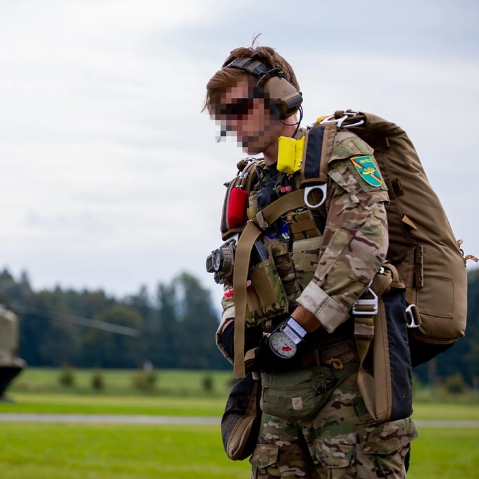 A person in military gear, including a backpack and helmet, stands on a grassy field with trees in the background, focused and prepared.