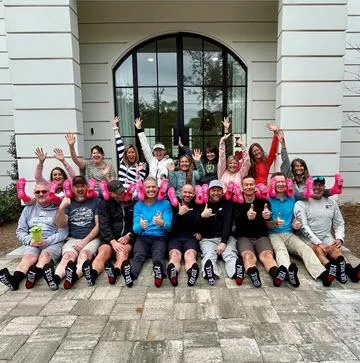 Large group of people sitting on a paved courtyard, raising their hands and holding pink dumbbells, all wearing matching black socks with white text, symbolizing team spirit, fitness, and community bonding.