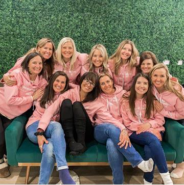 Group of ten women wearing matching pink hoodies, smiling and posing together on a teal couch against a green backdrop, representing teamwork, community, and empowerment.