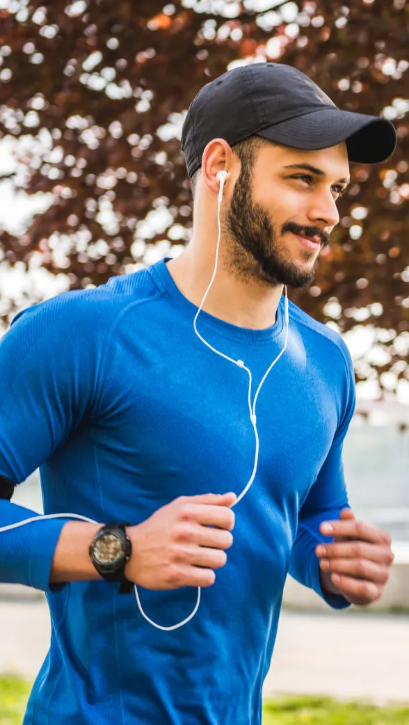 Young man jogging outdoors in a blue athletic shirt and black cap, smiling while listening to music with wired earbuds