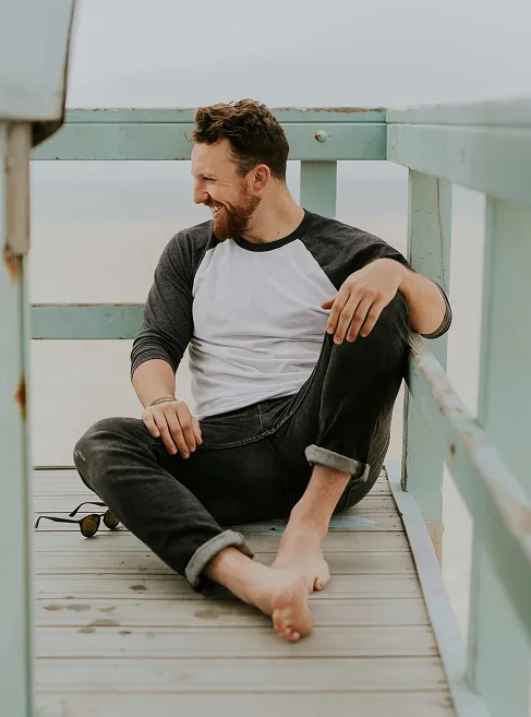 Relaxed man sitting barefoot on a wooden beach lifeguard stand, smiling and looking to the side, wearing a baseball-style shirt and jeans