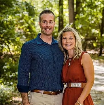  Smiling couple posing outdoors on a scenic wooded trail, dressed in smart casual attire, symbolizing partnership, success, and a balanced lifestyle.