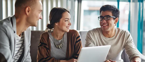 Three professionals engaged in a positive and collaborative discussion, working together on a laptop in a modern workspace, representing teamwork and digital learning within the Impact Nation Training Portal.