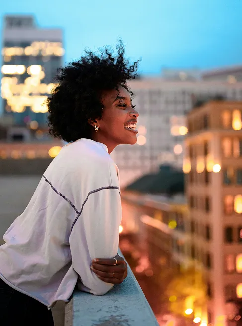 Smiling woman with curly hair leaning on a rooftop ledge at sunset, overlooking a brightly lit city skyline