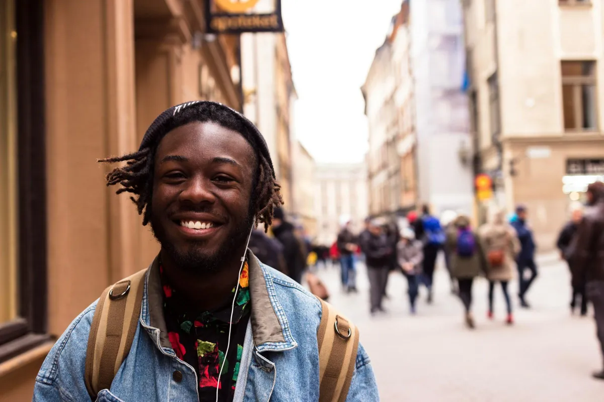 black man with locs in a jean jacket smiling near a building
