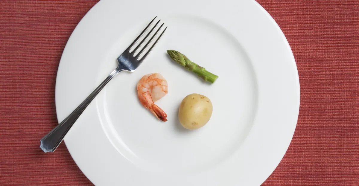 A white dinner plate with a fork holds a very small portion of food consisting of one shrimp, one baby potato, and one asparagus spear on a red textured placemat, illustrating restrictive eating, calorie restriction, or undereating.