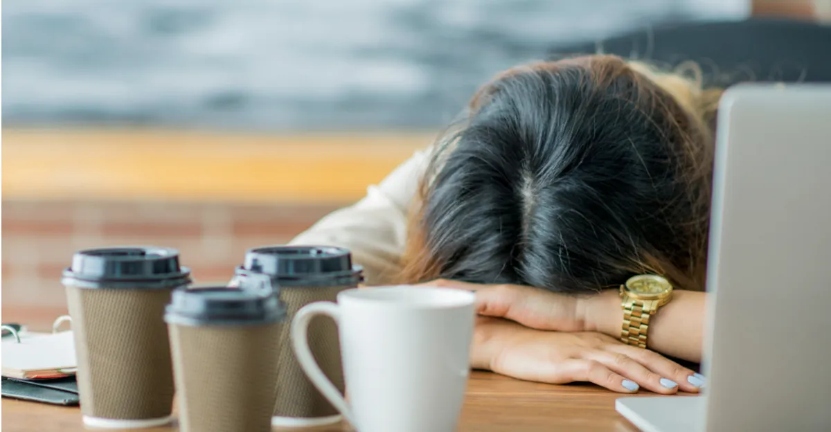 Woman resting her head on her arms at a desk beside coffee cups and a laptop, representing fatigue, brain fog, and low energy.