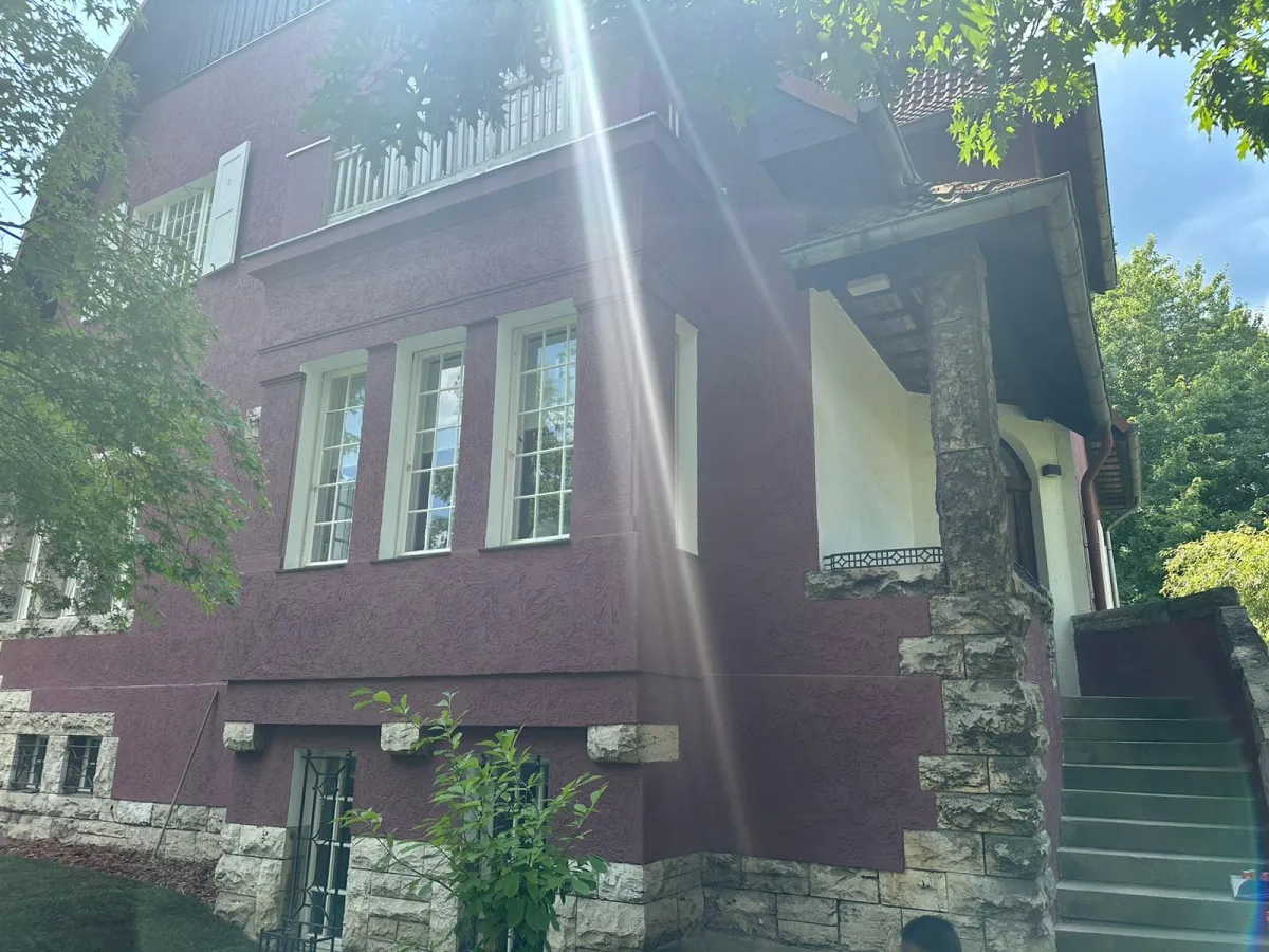 Außenansicht einer Villa mit dunkelvioletter Fassade, Natursteinsockel und weiß gerahmten Fenstern mit Balkon; starker Lens-Flare