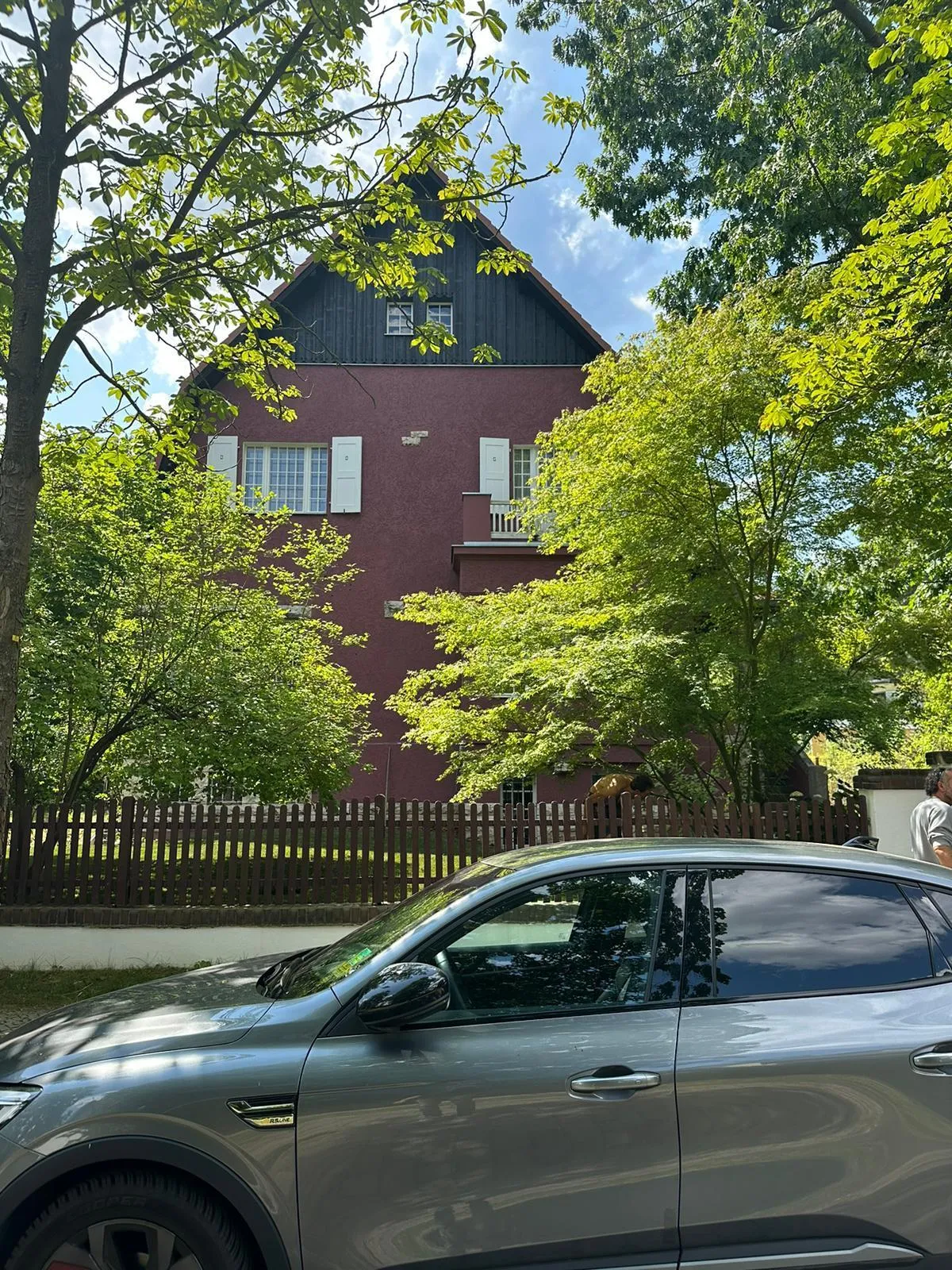 Außenaufnahme eines weinroten mehrstöckigen Wohnhauses; im Vordergrund parkt ein silberner SUV vor Zaun und Bäumen