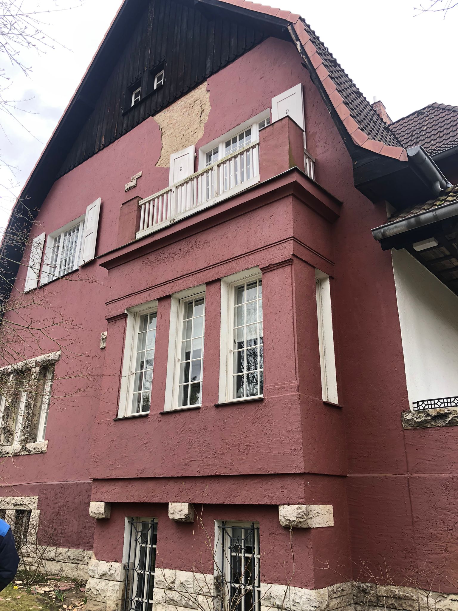 Seitenansicht eines altrosa verputzten Hauses mit Erker, weißem Balkongeländer und dunklem Holzgiebel; an der Fassade blättert teilweise der Putz ab.