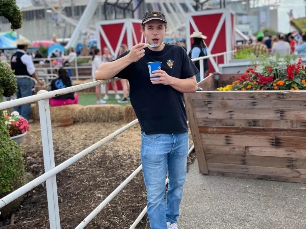 Young man in casual jeans and black t‑shirt leaning on a white railing at an outdoor fair, holding a blue drink cup with colorful flowers and barn-style booths in the background.