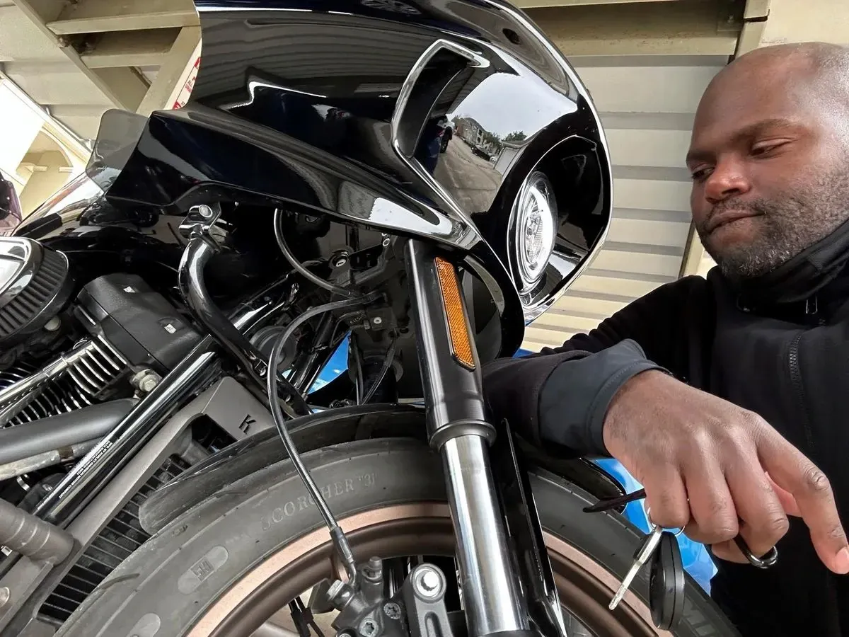 Close-up of a rider’s hand next to the front wheel and fairing of a black touring motorcycle, showing the headlight, suspension, and brake components during a maintenance check.