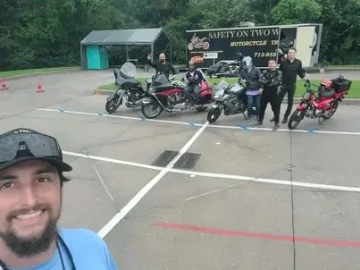 Group of motorcycle students and instructors posing with a mix of training bikes on a marked practice range in front of a “Safety On Two Wheels” trailer, celebrating after class.