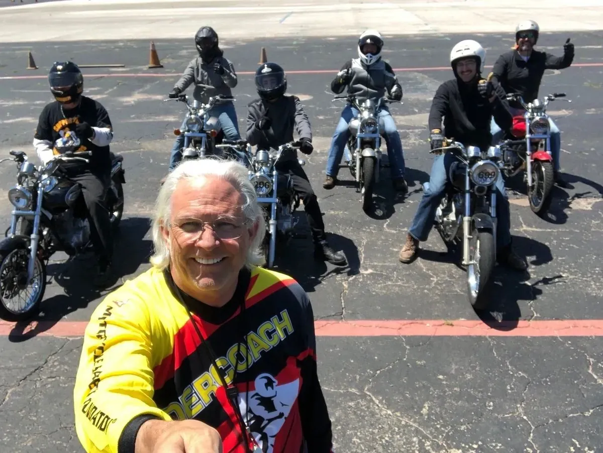 Motorcycle safety coach taking a selfie in front of a row of beginner riders sitting on training bikes in a parking-lot range, all wearing helmets and giving thumbs-up after class.