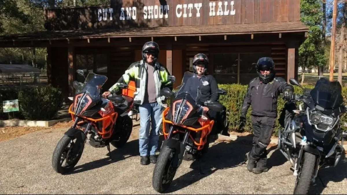Three adventure motorcyclists in full riding gear posing with two orange KTM adventure bikes and a black touring motorcycle in front of the rustic Cut and Shoot City Hall building.