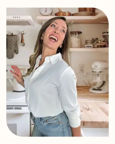 Smiling woman in a white shirt standing in a bright kitchen with baking tools and shelves in the background.