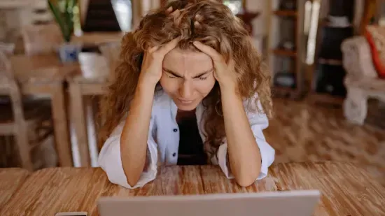 Frustrated woman sitting at a table with a laptop, holding her head in stress or overwhelm.