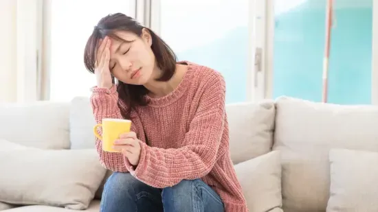 Woman sitting on a couch holding a yellow mug, touching her forehead with eyes closed, appearing tired or unwell.