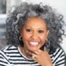 Smiling woman with voluminous curly gray hair wearing a black and white striped top, resting her chin on her hand.