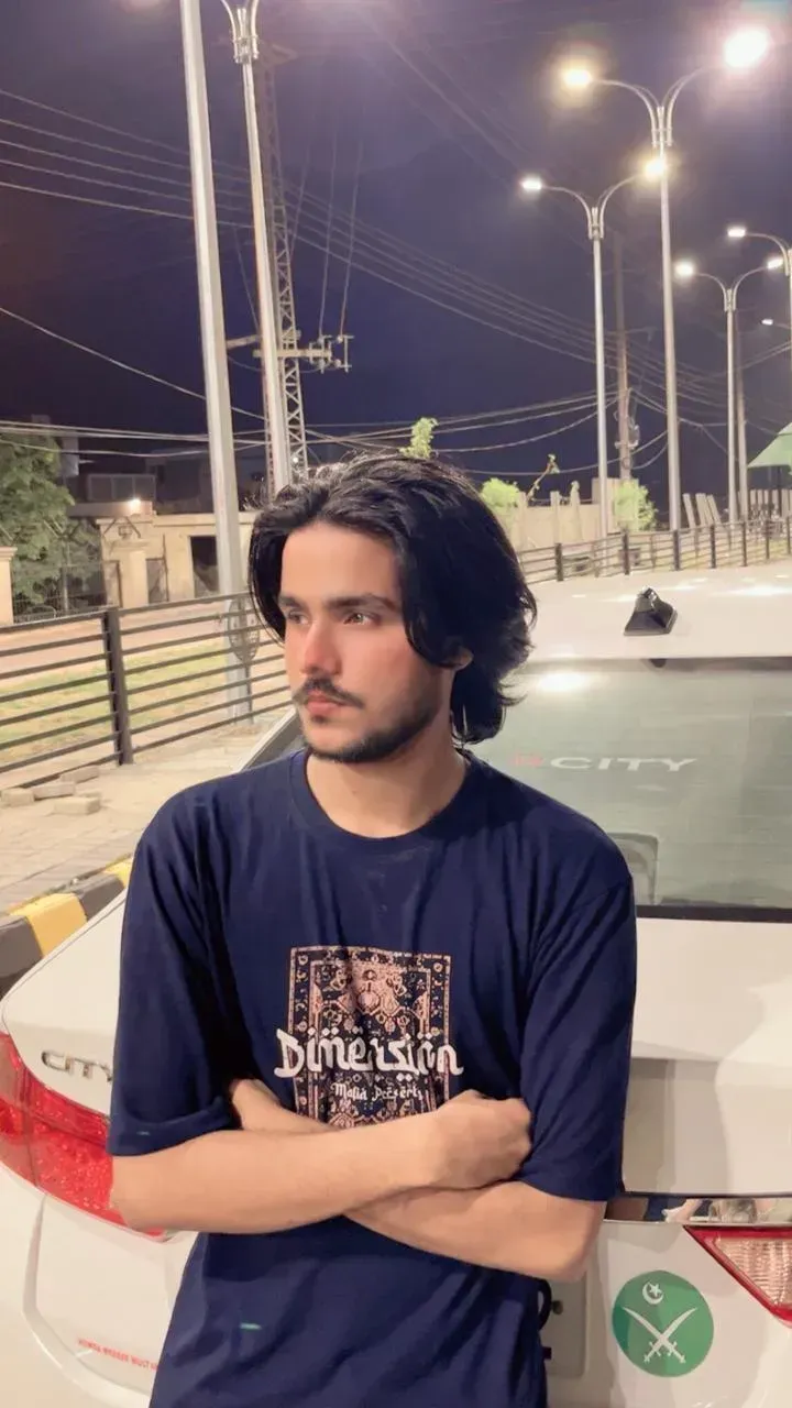 Portrait of a man with long hair looking thoughtfully to the left, standing outside on a city street at dusk, warm lighting.