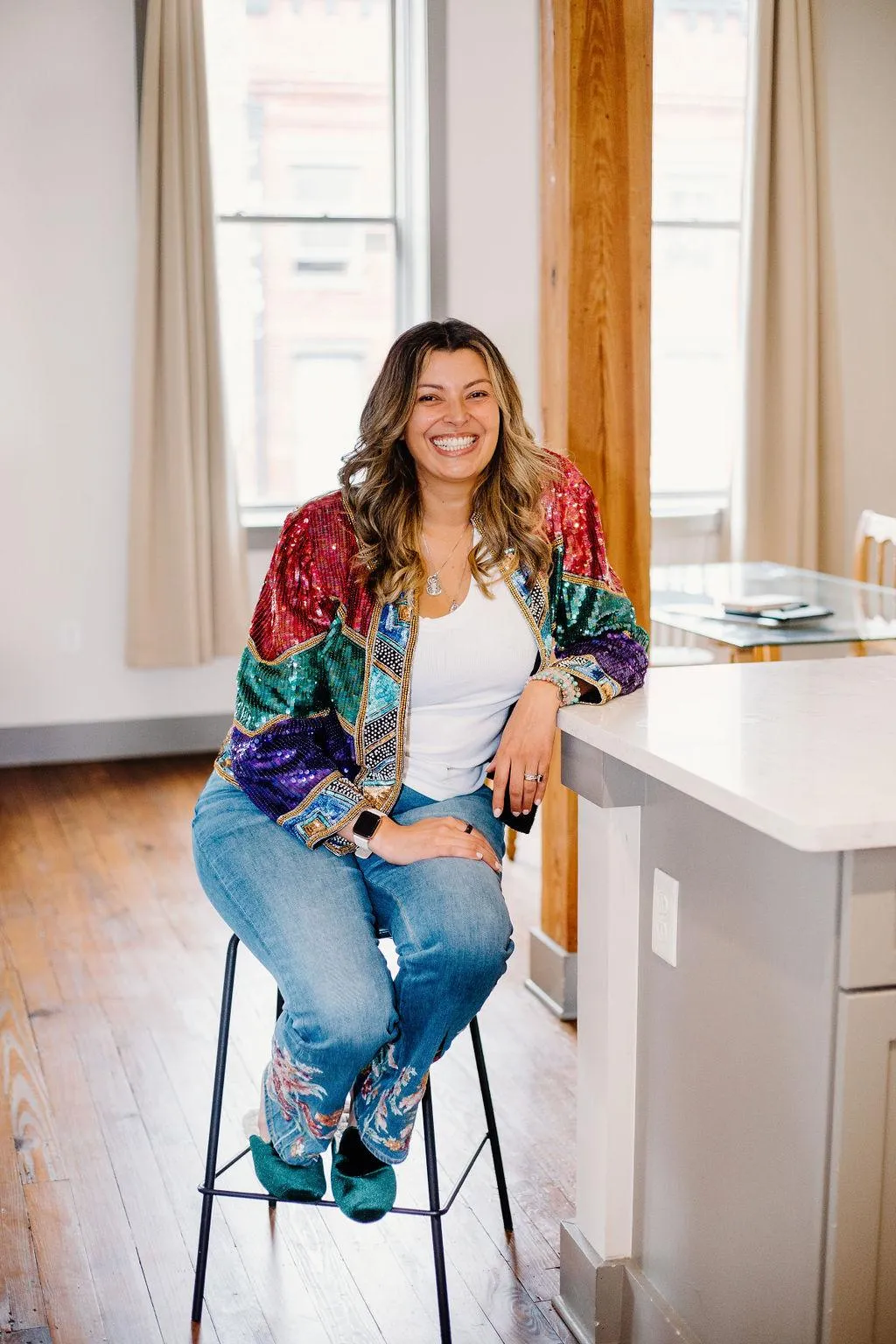 Gigi seating on a bar stool, smiling. Wearing jeans, a white t-shirt and a sequin jacket.