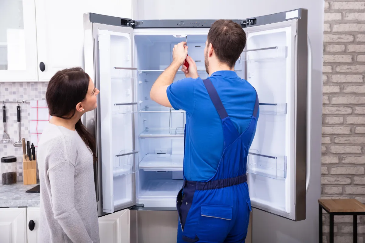 A technician repairing a refrigerator while a homeowner watches, representing solutions for loud refrigerator noise issues.