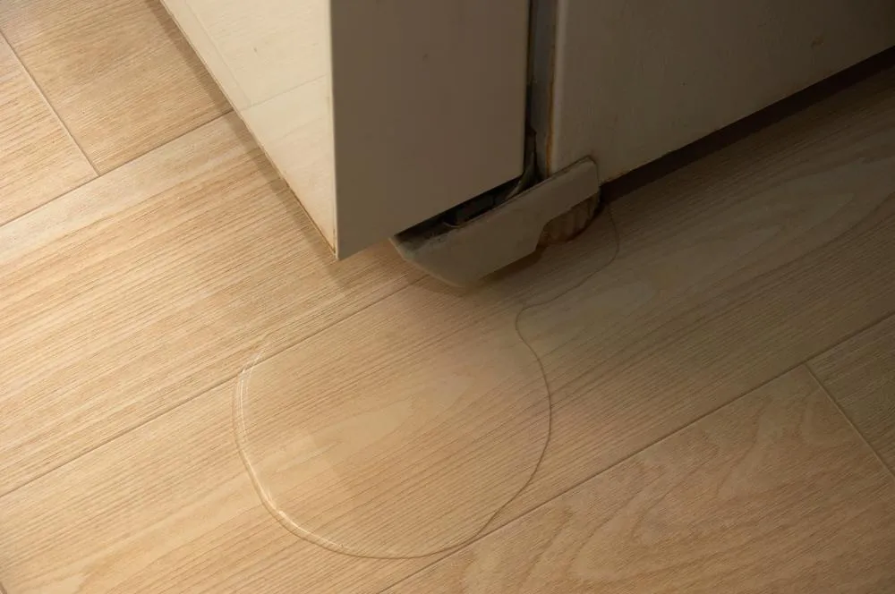 Water leaking from the bottom of a refrigerator onto a wooden kitchen floor, illustrating a common fridge leak issue.