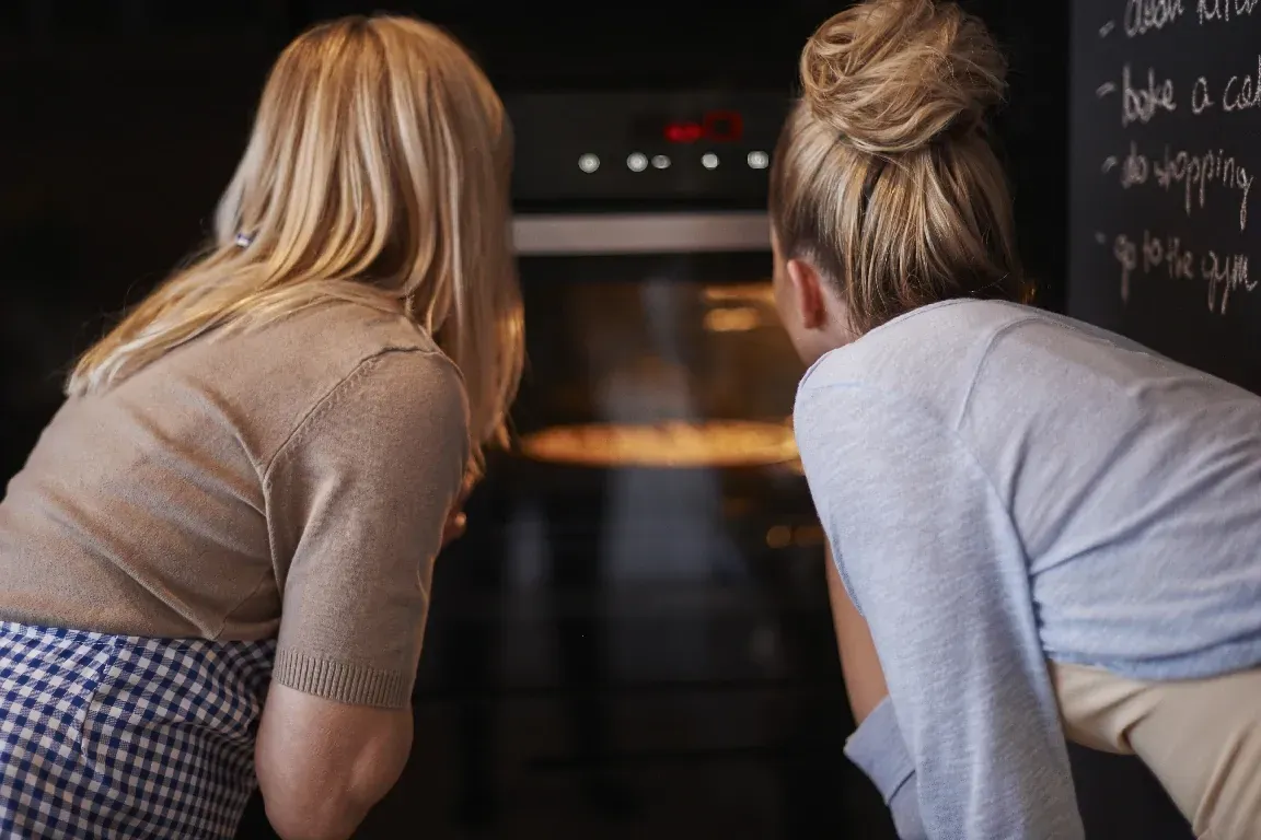 Two women are watching food bake inside an oven, representing troubleshooting common oven error codes.
