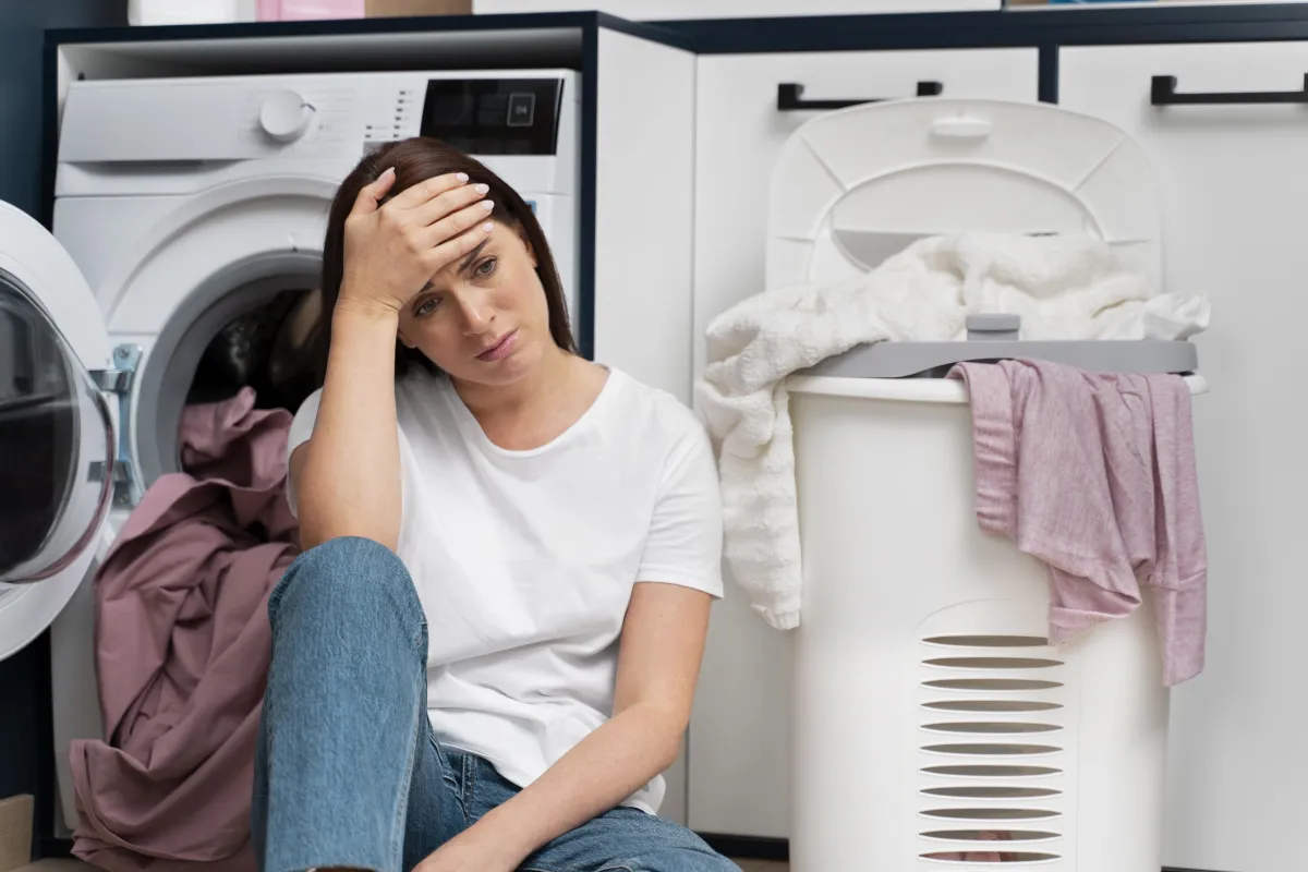 A woman sitting stressed beside a washing machine and a laundry basket, symbolizing dryer malfunction concerns.