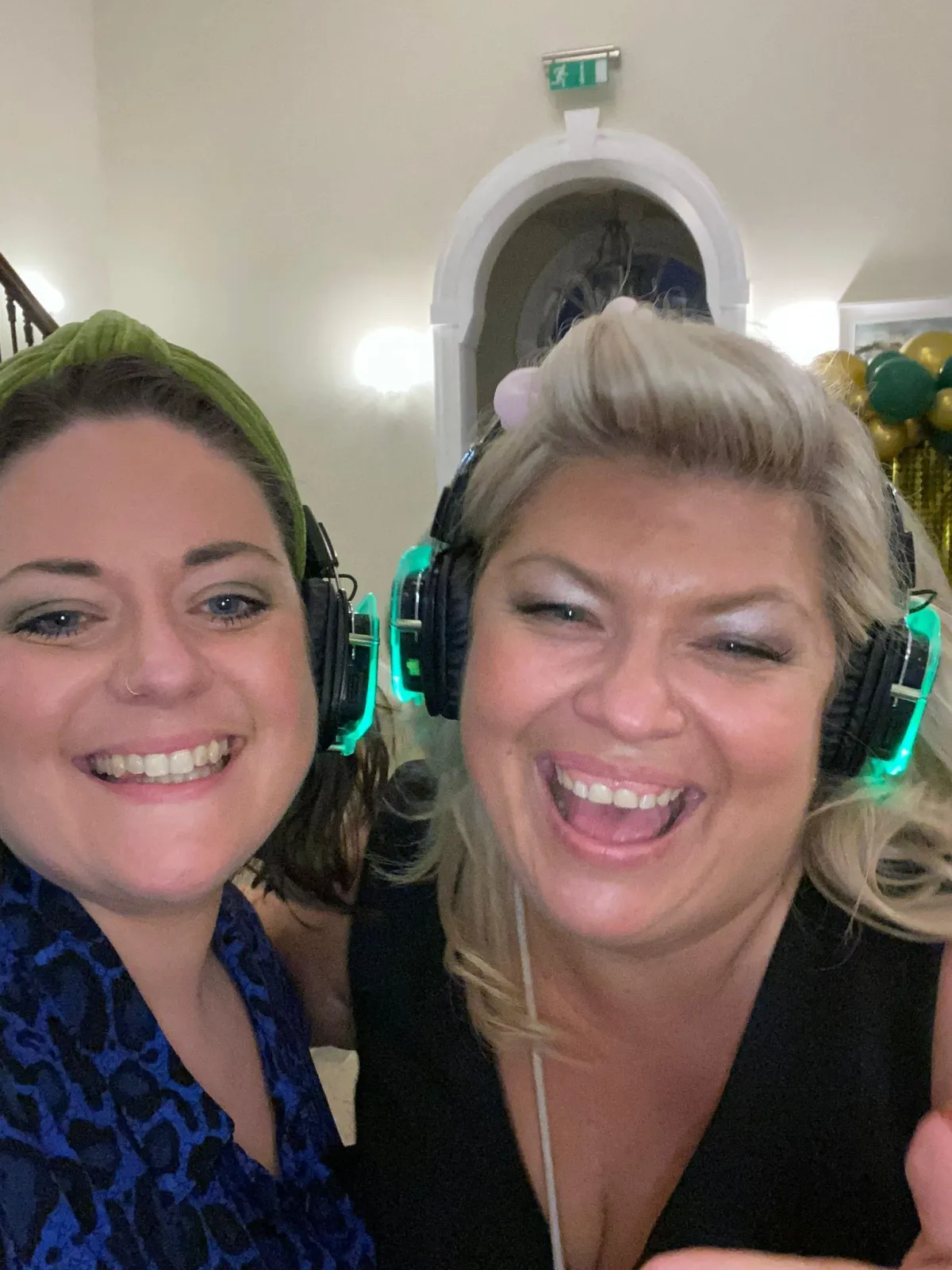 2 cis gendered women, one blonde with a sassy smile and one brunette smile and laugh at the camera for a selfie wearing their silent disco headphones at The Welly Boot Ball 2024