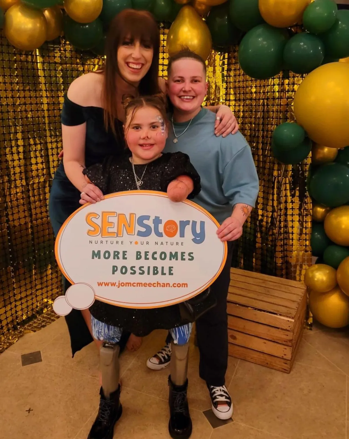 Founder and Director of senstory hq cic Jo McMeechan stands with her partner in front of the well boot ball photo wall holding branding. A little girl with limb difference wearing prosthetics smiles broadly at the camera at the front