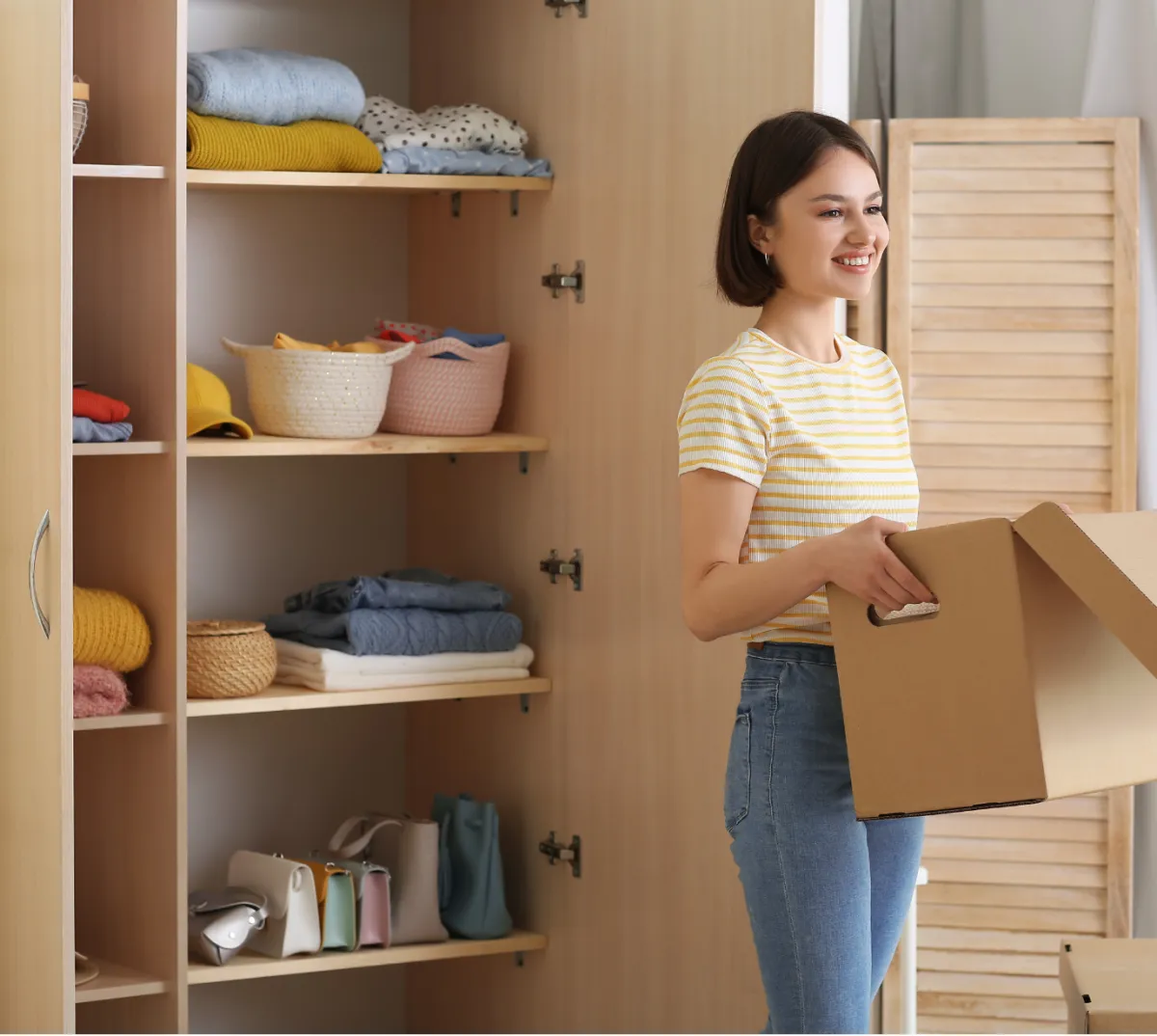 A woman in her room holds a cardboard box, suggesting she is preparing to unpack or organize items.