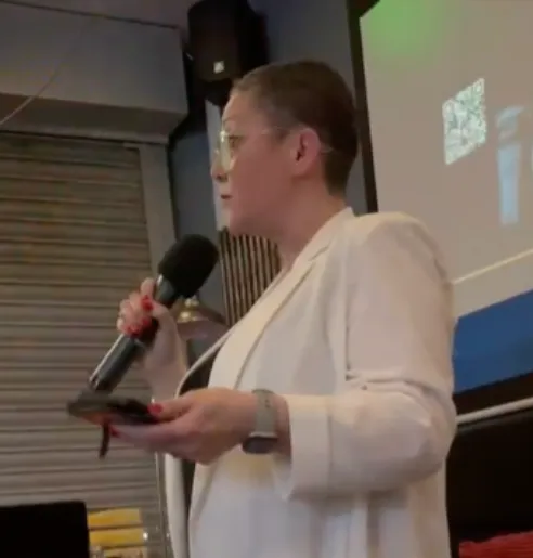 Sue Davies wearing white blazer, holding microphone. Speaking at an event.