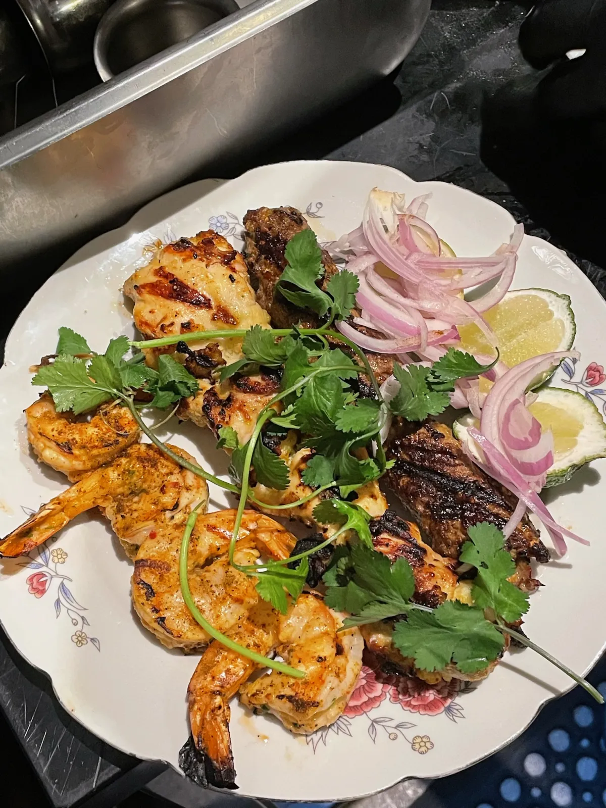 A decorative plate filled with grilled skewers, seasoned shrimp, sliced red onions, fresh cilantro, and lime wedges.