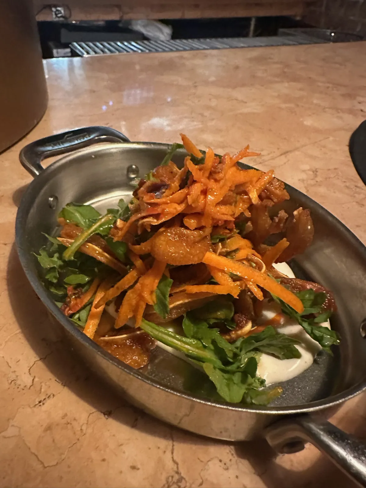 A small metal serving dish with handles containing a layered salad with dark greens, shredded orange carrots or vegetables, and a creamy white base on a warm-toned countertop.