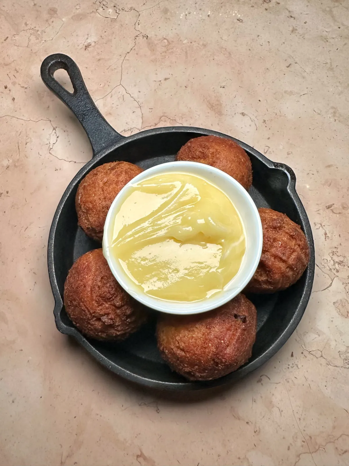 An overhead view of several golden-brown round fritters or hushpuppies arranged in a black cast-iron skillet, with a small white bowl of yellow butter or sauce placed centrally on top, all set against a textured light-toned countertop.
