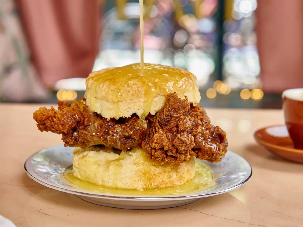 A crispy fried chicken sandwich on a biscuit, sitting on a decorative plate, while syrup is being poured over the top from above.