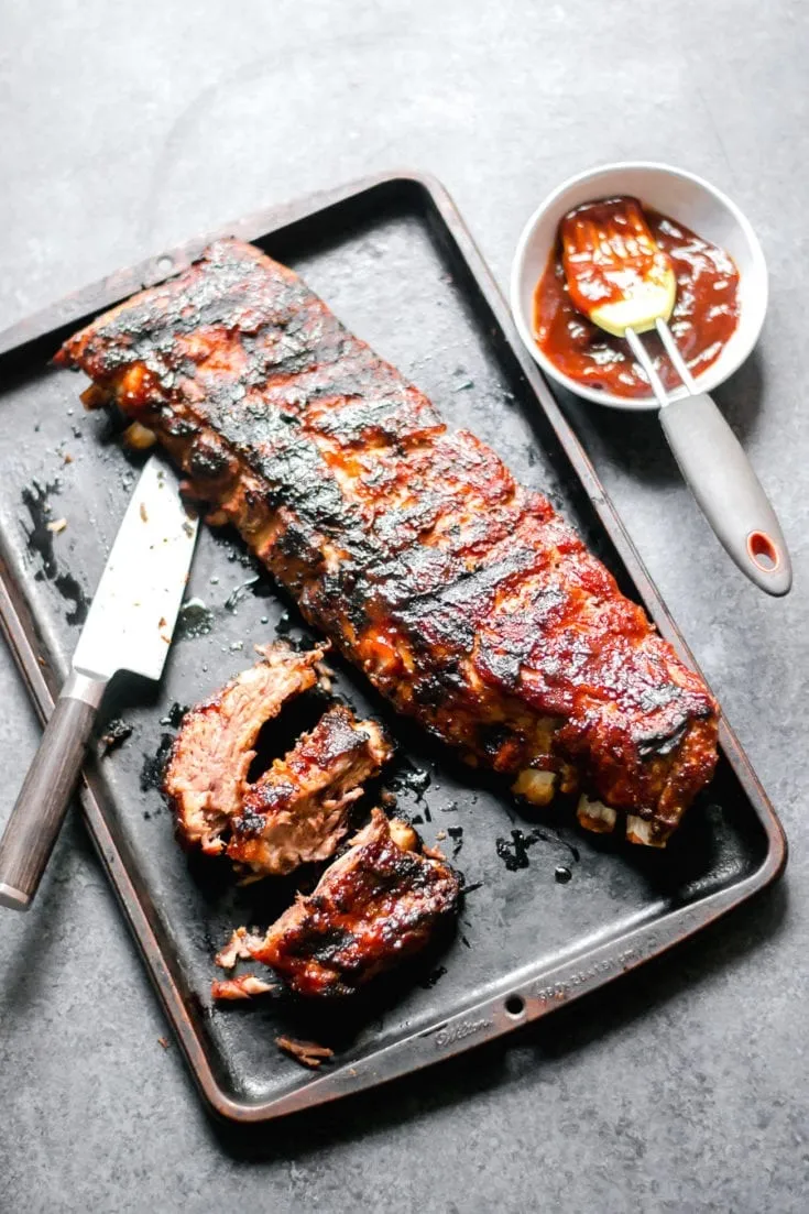 A rack of barbecued ribs with a charred, glazed exterior rests on a dark baking sheet, with a few cut pieces, a knife, and a small bowl of barbecue sauce with a basting brush nearby.