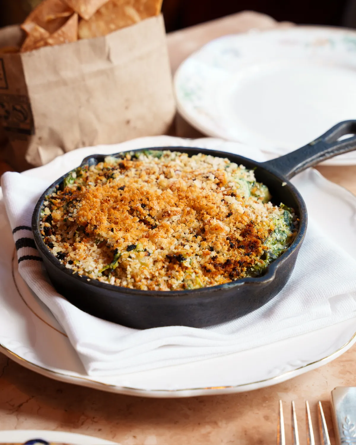 A small black cast-iron skillet filled with a baked, creamy green dip topped with a golden-brown breadcrumb crust, resting on a white napkin and plate next to a brown paper bag of chips.