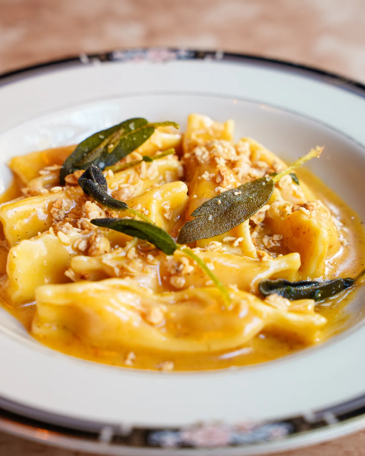 A close-up shot of folded pasta in a rich, glossy sauce, garnished with crispy fried sage leaves and a sprinkle of crushed nuts or breadcrumbs, served in a decorative white plate.