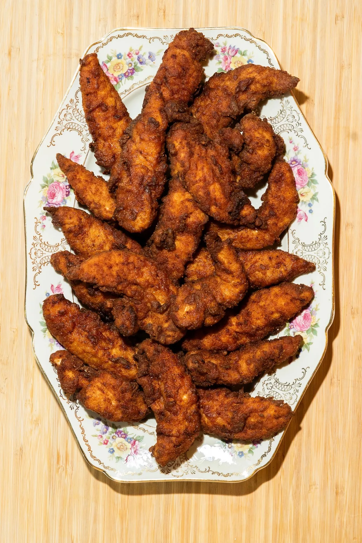 An overhead view of a decorative white platter with a floral pattern, piled high with golden-brown, crispy fried chicken tenders resting on a wooden surface.