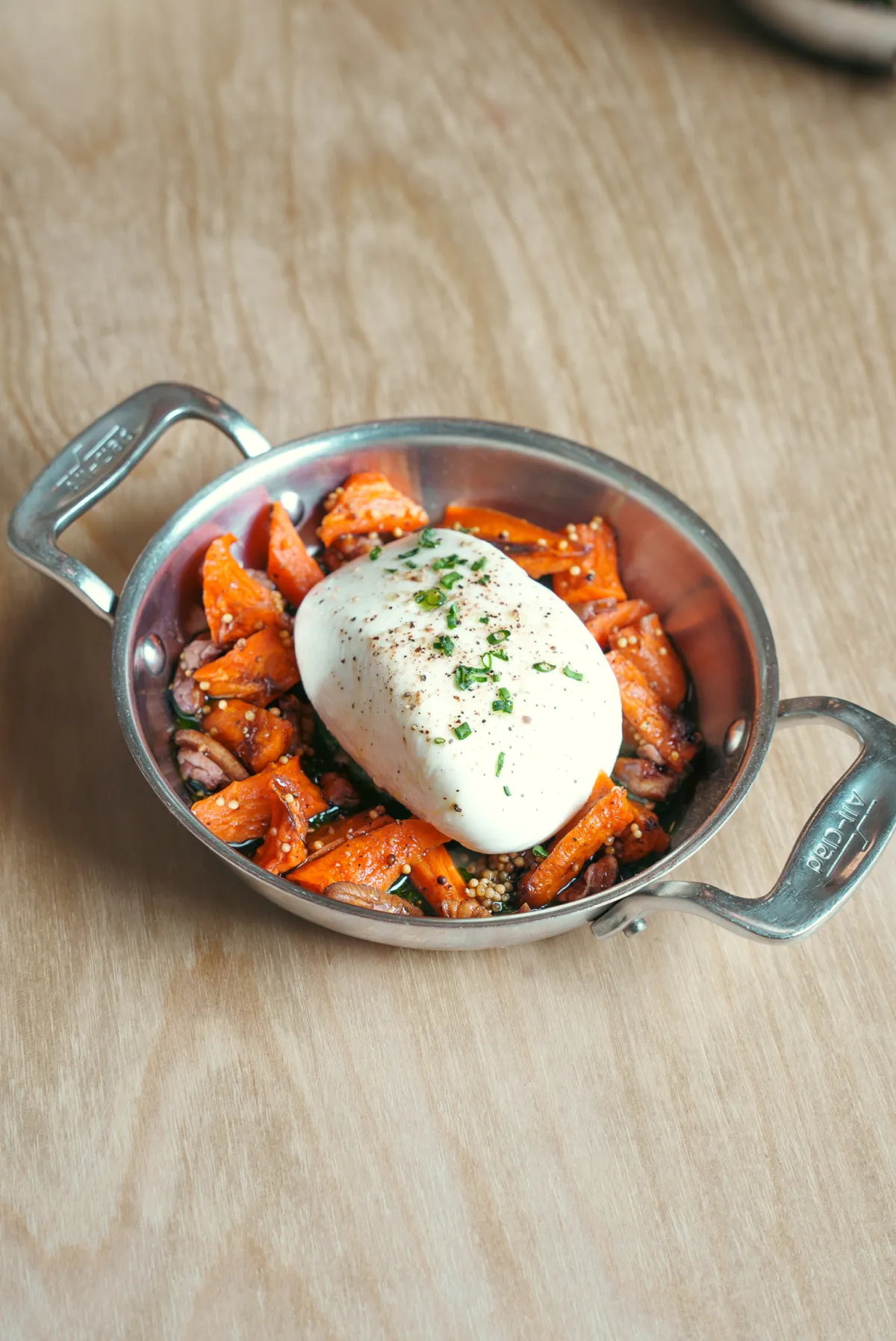 A small metal serving dish with handles rests on a wooden surface, containing roasted orange carrots and mustard seeds topped with a large, creamy white ball of burrata cheese sprinkled with herbs and pepper.